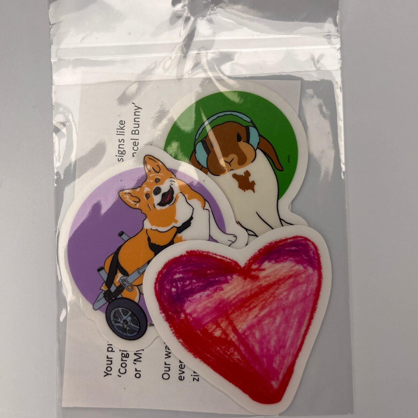 Three GoodOnU vinyl stickers a corgi on a purple background with wheels, a bunny wearing noise cancelling headphones on a green background and a my heart sticker.