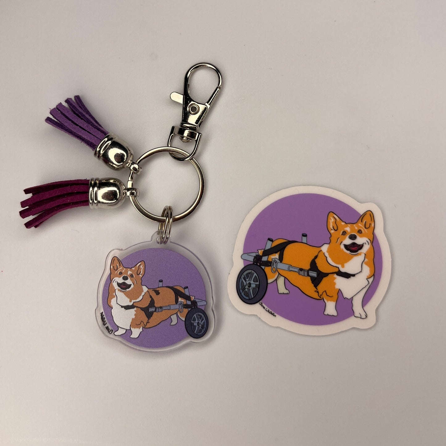 Corgi charm with two tassels metal lobster clasp and keyring next to a 2" x 2" vinyl Corgi sticker on a purple background 