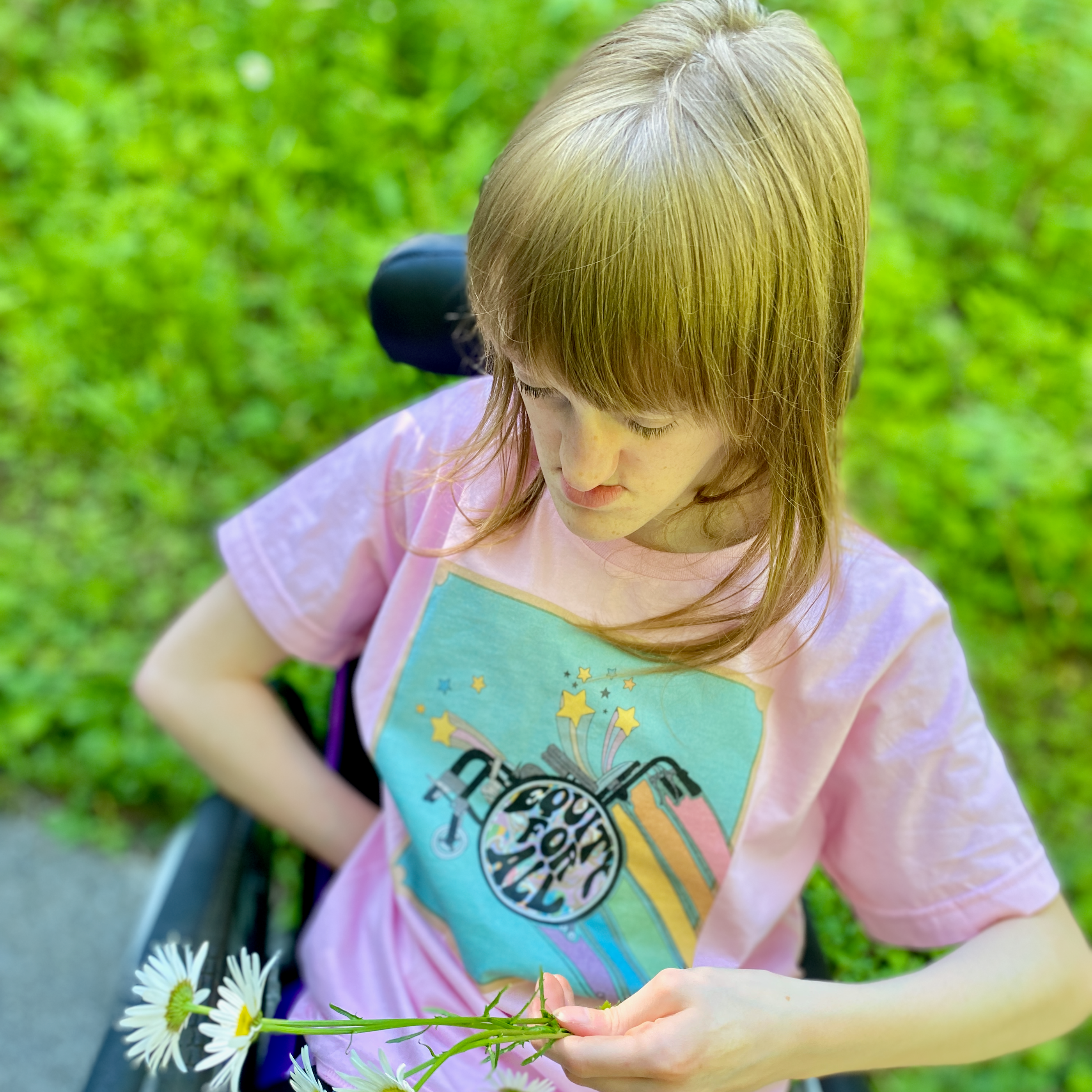 Teen holding daisy bouquet seated in wheelchair with grass background wearing cool and colourful t-shirt of flying wheelchair on blue background with shooting stars trailing rainbow and slogan Equity for All